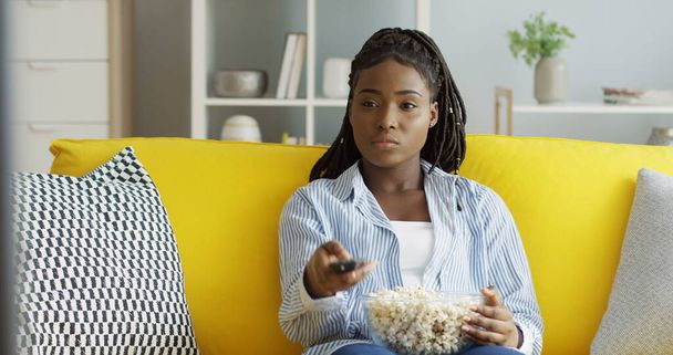 Όμορφη Αφροαμερικανή νεαρή γυναίκα που βλέπει τηλεόραση με ένα μεγάλο μπολ ποπ κορν, αλλάζει κανάλια και κάθεται στον κίτρινο καναπέ στο μοντέρνο σαλόνι. Εσωτερικά - Φωτογραφία, εικόνα