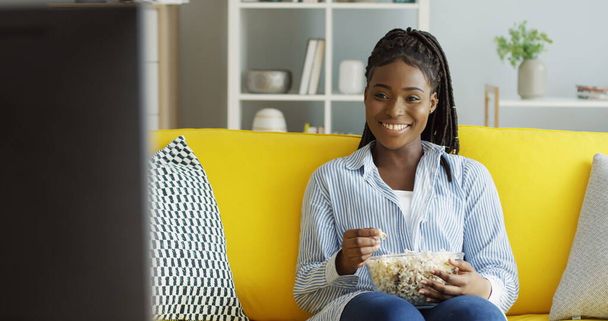 Mooie Afro-Amerikaanse jonge vrouw in het blauwe shirt die popcorn eet terwijl ze op tv komedie kijkt en thuis op de gele bank lacht. Binnenkant - Foto, afbeelding