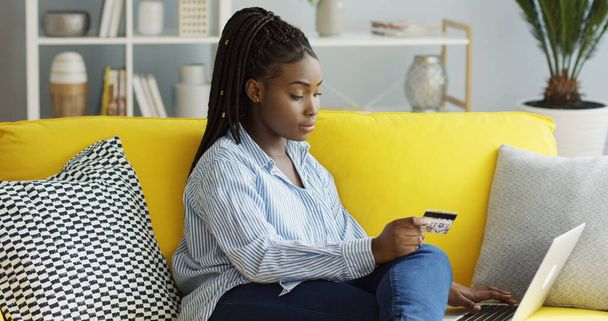Jovem mulher afro-americana atraente fazendo compras on-line ao inserir um número de cartão de crédito no computador portátil em casa. Para dentro
 - Foto, Imagem