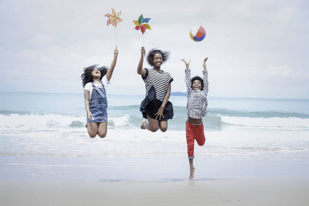 Cute groups of kids have fun on sandy summer beach with blue sea, happy childhood friends  jump together and play on tropical beach,  children raise hand up, hold ball and paper windmill - Photo, Image