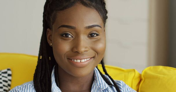 Gros plan de la jolie jeune femme afro-américaine avec une queue de cochons regardant et posant devant la caméra sur le canapé jaune. Intérieur - Photo, image