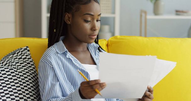 Close up da mulher afro-americana segurando folhas de papel e lápis nas mãos e verificando documentos no sofá em casa. Interior
 - Foto, Imagem