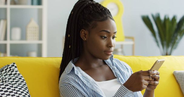 Primer plano de la joven atractiva mujer afroamericana con una cola desplazándose y grabando en el teléfono inteligente mientras está sentada en el sofá en la acogedora sala de estar. De interior
 - Foto, imagen