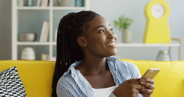 Primer plano de la joven atractiva mujer afroamericana con una cola desplazándose y grabando en el teléfono inteligente mientras está sentada en el sofá en la acogedora sala de estar. De interior
 - Foto, Imagen