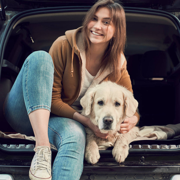Close up of woman in blue jeans next to golden retriever sitting in open trunk of black car - Photo, Image