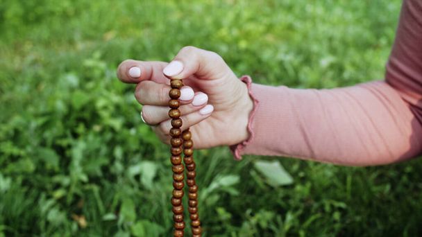 Woman lit hand close up counts rosary - malas strands of gemstones beads used for keeping count during mantra meditations. Girl sits on summer nature. - Photo, image