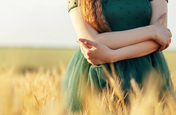 Beautiful woman in green dress walking in field and touches ears of wheat with hand at sunset light, girl enjoying summer nature landscape - Foto, Imagen