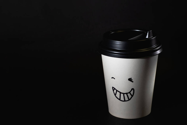 one white disposable coffee cup closed with a black lid with a smile on the body, stands on a black background, isolated on black. - Valokuva, kuva