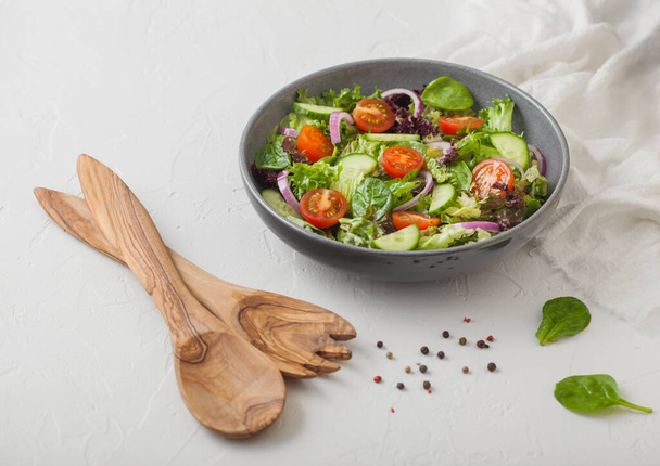 Healthy fresh vegetarian vegetables salad with lettuce and tomatoes, red onion and spinach in grey bowl plate on white table background with spatula fork and and spoon. - Foto, Bild