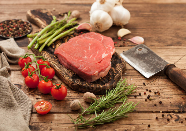 Fresh raw barbeque braising beef steak on chopping board with asparagus and garlic with cherry tomatoes and salt with pepper on wooden background with hatchet. - Foto, Imagem