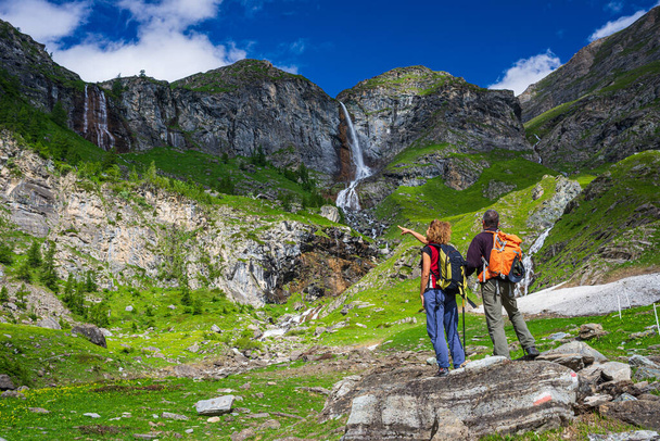 Ein paar Wanderer betrachten den Wasserfall auf dem Berg. Zwei Personen mit Rucksack Outdoor-Aktivität in malerischer alpiner Landschaft, Sommerurlaub in den Alpen, Fitness-Wellness-Freiheit - Foto, Bild