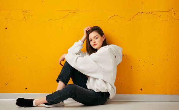 Young pretty calm relaxing girl in a white hoodie poses sitting on the floor against a yellow concrete wall. - Foto, imagen