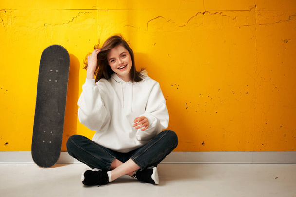 Young pretty happy smiling girl in a white hoodie with a skateboard poses against a yellow concrete wall. - Φωτογραφία, εικόνα