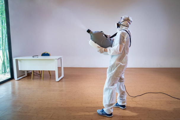 Professional technical man in prevention suit sprays sterilising solution by electrical spray machine on wood floor and white background with studio light. - Photo, Image