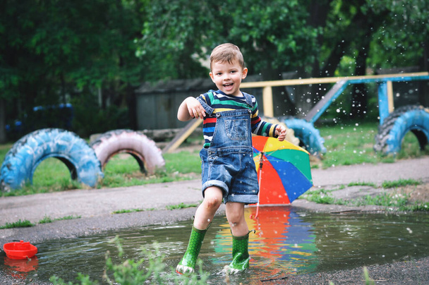 Μικρό χαριτωμένο αγόρι με τζιν φόρμες σορτς και πράσινες λαστιχένιες μπότες παίζει σε μια μεγάλη λακκούβα. Εκτοξεύει βάρκες και πηδάει σε μια λακκούβα.. - Φωτογραφία, εικόνα