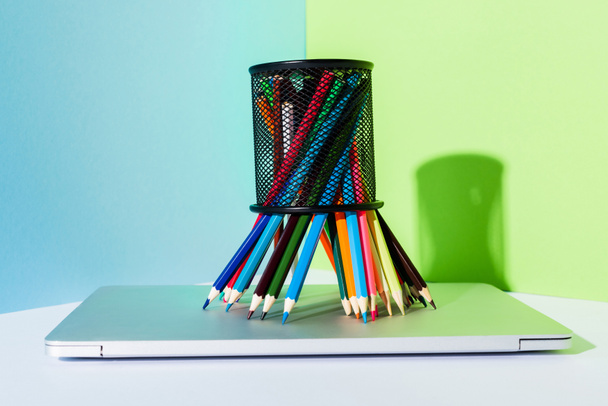 цветные карандаши в держателе карандаша на современном ноутбуке на синем, зеленом и белом фоне
 - Фото, изображение