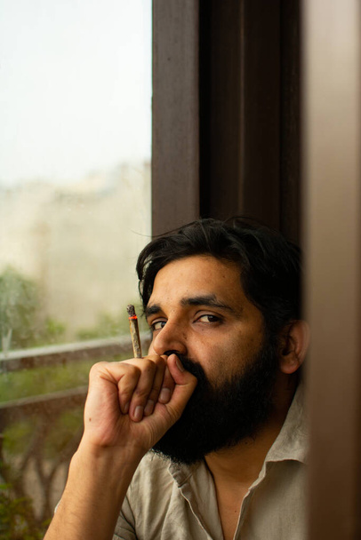 髭を生やしたインド人の男が紙の中にクルタ・ピジャマの喫煙用雑草を巻いていた。彼はカメラを見て家に座っている。背景に空を洗い流し - 写真・画像