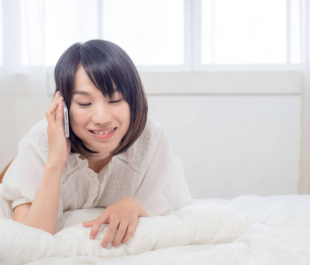 Portret pięknej młodej Azjatki rozmawiającej przez telefon komórkowy leżącej na łóżku w pokoju w domu. Styl życia z młodą dziewczyną i czas relaksu Concept. - Zdjęcie, obraz