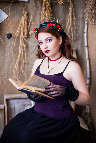 Portret van een jonge, aantrekkelijke heks zittend aan een tafel met magische boeken en rollen tegen een achtergrond van alchemische ingrediënten, kolven, flessen en gedroogde kruiden. - Foto, afbeelding