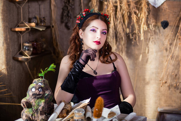 Portrait of a young, attractive witch sitting at a table with magic books and scrolls against a background of alchemical ingredients, flasks, bottles, and dried herbs. - 写真・画像