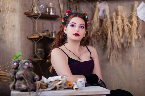 Portrait of a young, attractive witch sitting at a table with magic books and scrolls against a background of alchemical ingredients, flasks, bottles, and dried herbs. - Zdjęcie, obraz