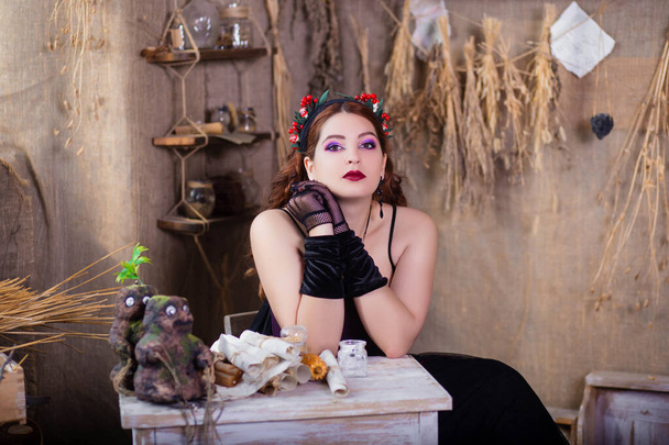 Ritratto di una giovane e attraente strega seduta a tavola con libri magici e pergamene su uno sfondo di ingredienti alchemici, fiaschi, bottiglie ed erbe essiccate. - Foto, immagini