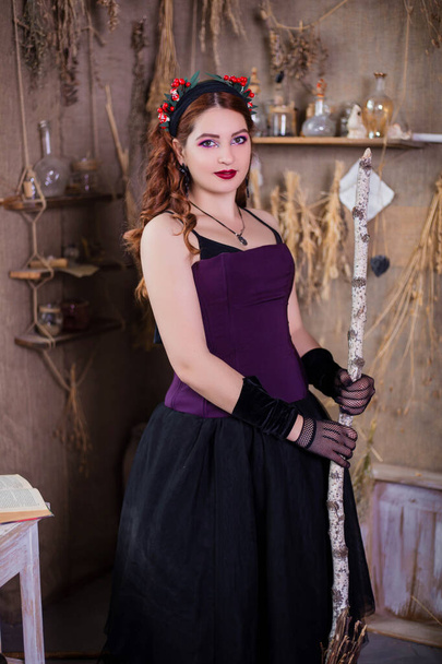 A young, attractive witch with a broom in her hands is standing against a background of alchemical ingredients, flasks, bottles and dried herbs. - Foto, immagini