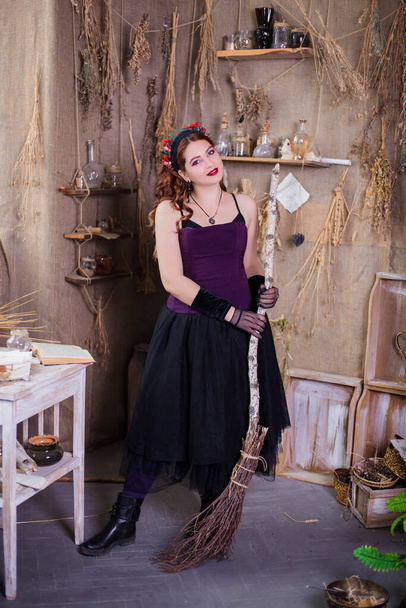 A young, attractive witch with a broom in her hands is standing against a background of alchemical ingredients, flasks, bottles and dried herbs. - Photo, Image