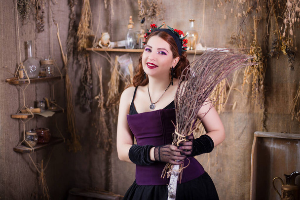 A young, attractive witch with a broom in her hands is standing against a background of alchemical ingredients, flasks, bottles and dried herbs. - Fotografie, Obrázek