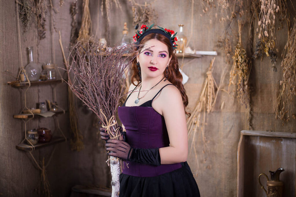 Een jonge, aantrekkelijke heks met een bezem in haar handen staat tegen een achtergrond van alchemische ingrediënten, kolven, flessen en gedroogde kruiden. - Foto, afbeelding