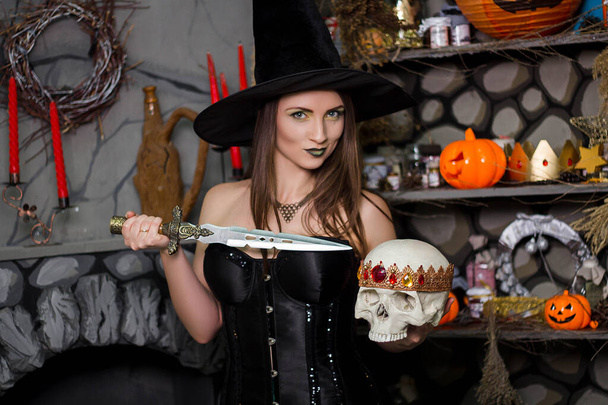 A young, beautiful woman in a green skirt, a black corset and a black hat holds a knife and a skull with a crown in her hands. Witch costume, Halloween, photo studio, model with clean skin. - Foto, Imagen