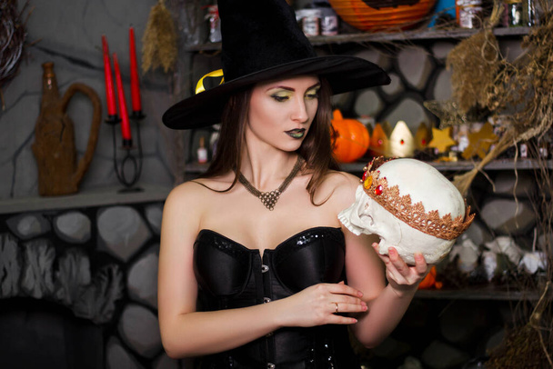 A young, beautiful woman in a green skirt, a black corset and a black hat holds a skull with a crown in her hands. Witch costume, Halloween, photo studio, model with clean skin. - Photo, image