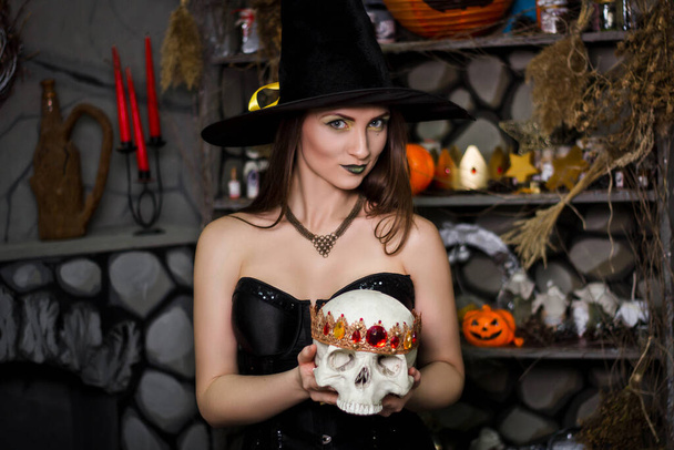 A young, beautiful woman in a green skirt, a black corset and a black hat holds a skull with a crown in her hands. Witch costume, Halloween, photo studio, model with clean skin. - Фото, зображення