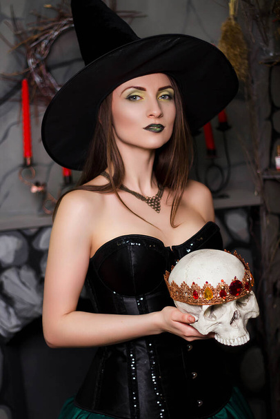 A young, beautiful woman in a green skirt, a black corset and a black hat holds a skull with a crown in her hands. Witch costume, Halloween, photo studio, model with clean skin. - Fotó, kép