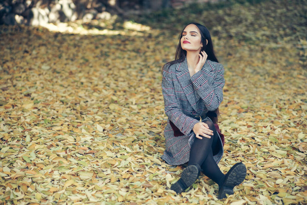 Beautiful girl wearing winter coat sitting on the floor of an urban park full of autumn leaves. - Photo, Image