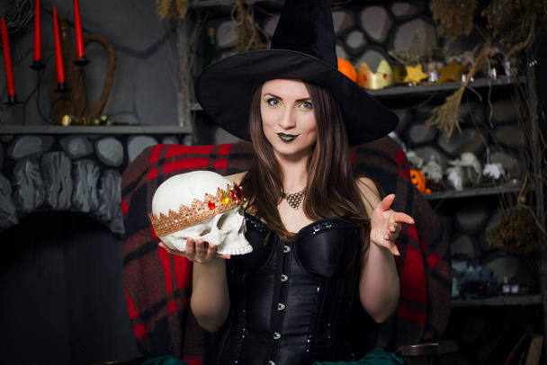 A young, beautiful witch in a black pointed hat and corset holds a skull in his hands and conjures seated on a armchair.  - Foto, afbeelding