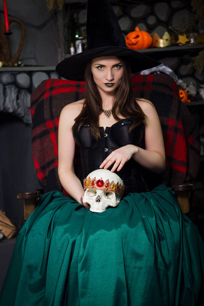 A young, beautiful witch in a black pointed hat and corset holds a skull in his hands and conjures seated on a armchair.  - Fotoğraf, Görsel