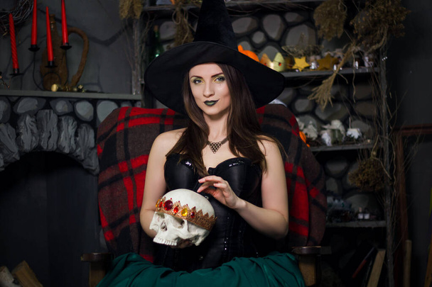 A young, beautiful witch in a black pointed hat and corset holds a skull in his hands and conjures seated on a armchair.  - Foto, Bild