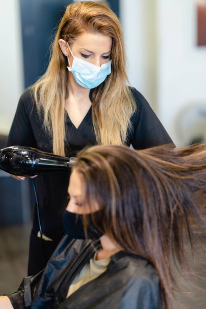 Fryzjer suszenie jej klientów włosy z suszarką do włosów noszenie masek ochronnych w centrum urody. - Zdjęcie, obraz