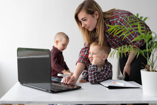 A schoolboy is doing quarantined home lessons. Distance learning. Online education of a child at home in a laptop. Webcam lessons. Mom helps her son with lessons. The second son calls his mother to play with him. Social distance. Place for text. - Photo, Image