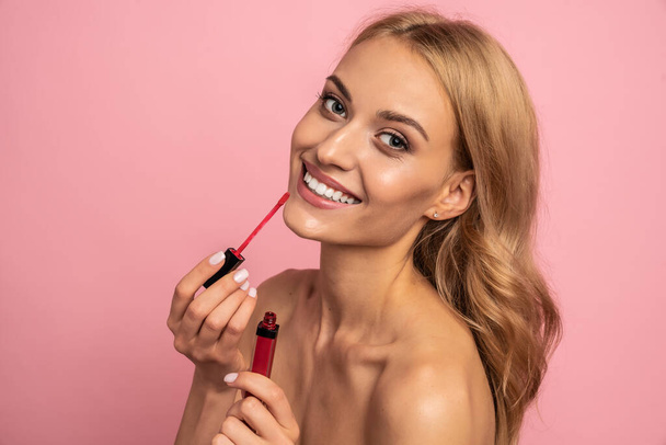 Portrait of cute, charming girl testing new lip gloss isolated on pink background, perfection, wellbeing advertisement concept. - Photo, Image