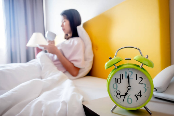 The girl sits in the morning drinking coffee on a bed covered with white linen. There is an alarm clock at 7:00 am in the bedroom. - Photo, Image