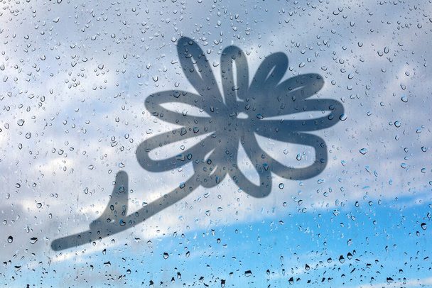 Квітковий малюнок на мокрій склянці. Скляне вікно з дощами на тлі блакитного неба. Емуляція дитячих малюнків
. - Фото, зображення