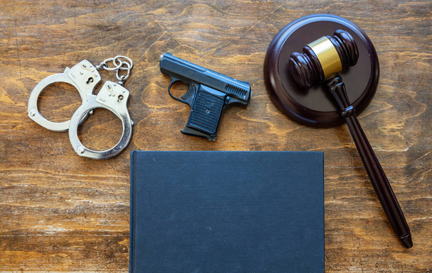 法律犯罪処罰の概念。裁判官の手荷物、拳銃、手錠、木製の机の背景にある空白のカバー法的本、トップビュー、コピースペース. - 写真・画像