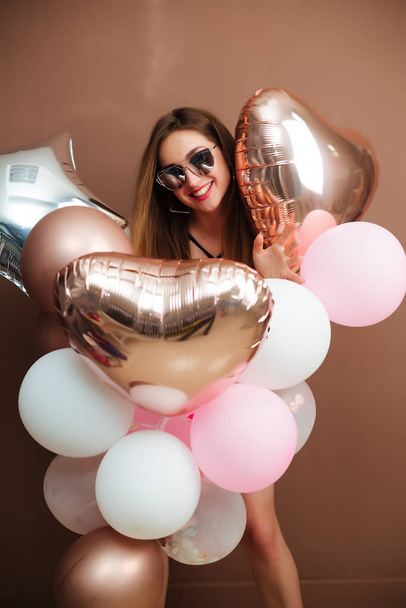 Slender girl in a bodysuit and colored balloons in her hands stands on a brown background in a festive mood posing in front of the camera. - Foto, afbeelding