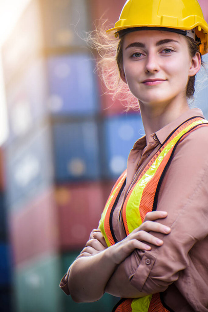 Νεαρή με αυτοπεποίθηση Καυκάσια γυναίκα μηχανικός φορώντας κίτρινο κράνος ασφαλείας και ελέγξτε για έλεγχο φόρτωσης εμπορευματοκιβωτίων κουτί από φορτηγό πλοίο για εισαγωγή και εξαγωγή - Φωτογραφία, εικόνα