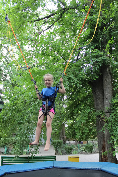 Το χαμογελαστό ευτυχισμένο κορίτσι άλμα με bungee στο τραμπολίνο στο πάρκο κατά πράσινα δέντρα - Φωτογραφία, εικόνα
