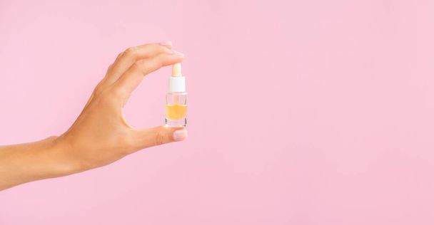 Beauty-Produkte Banner mit Kopierfläche. Tropfflasche mit Öl-Attrappe. Weibliche Hand hält gelbes Kosmetiköl in einem klaren Glas auf rosa Hintergrund - Foto, Bild