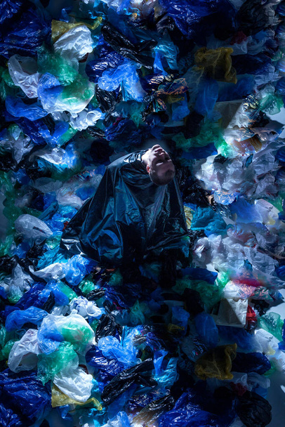 Vista superior de una imagen artística de un joven en el estudio, rodeado de bolsas de basura de plástico vacías. Contenido con problemas ecológicos, evidencia de la contaminación, aislado en la pared oscura del estudio
. - Foto, imagen