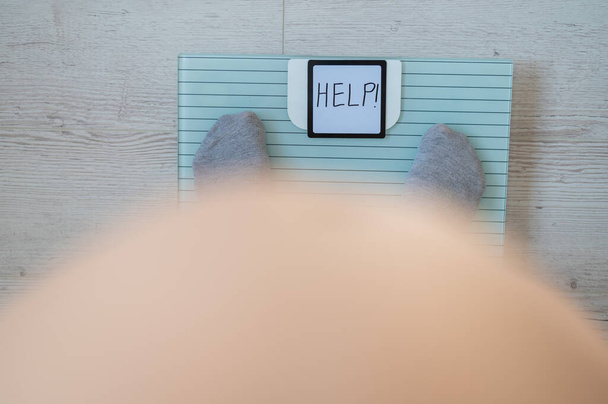 Faceless fat woman measures weight on a bathroom scale. Top view of a large naked female belly and feet in gray socks on the scales with the word help. The inscription on the screen help. Obesity. - Фото, изображение